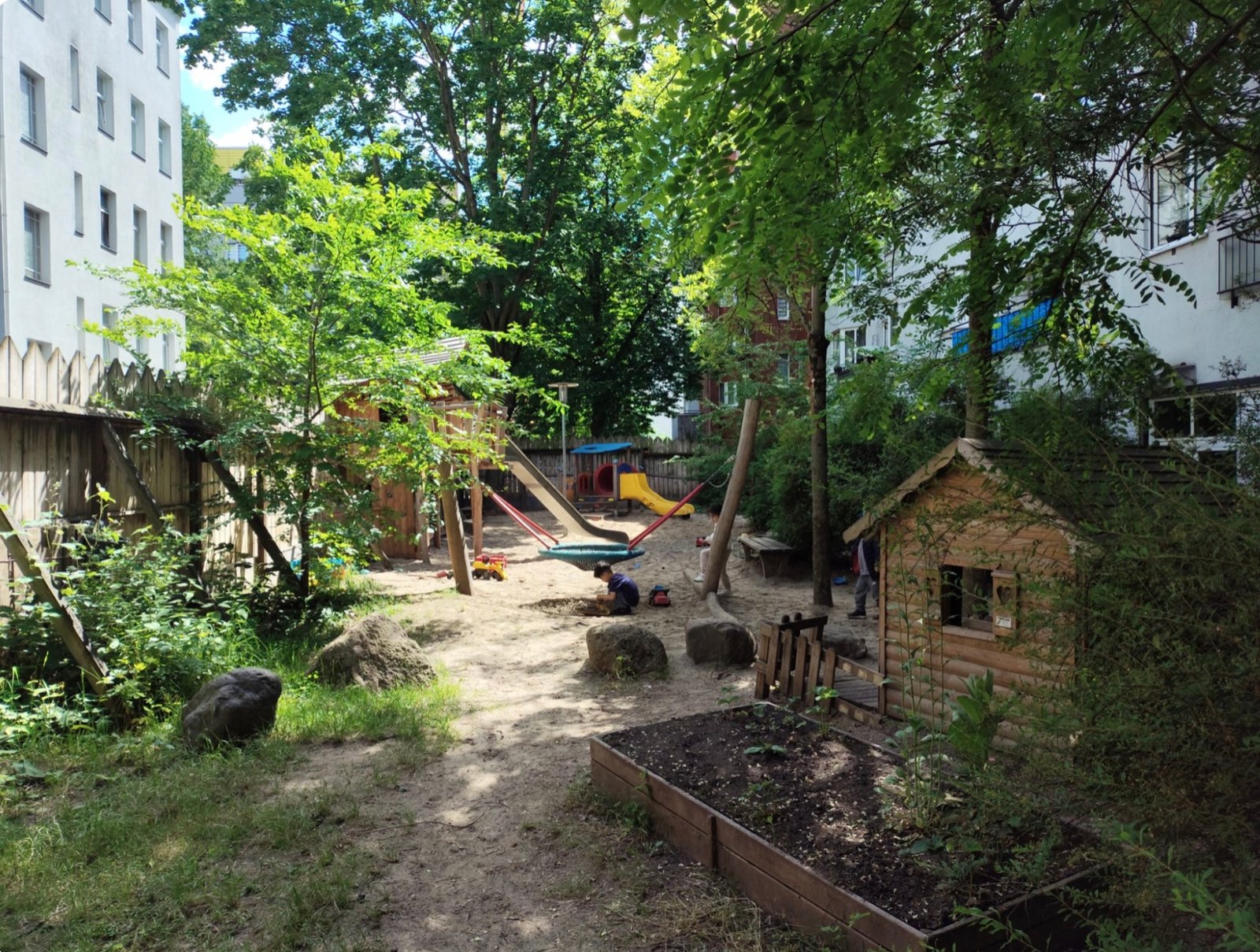 Begrünter Außenspielbereich mit Sandfläche, Spielgeräten und Holzhäuschen